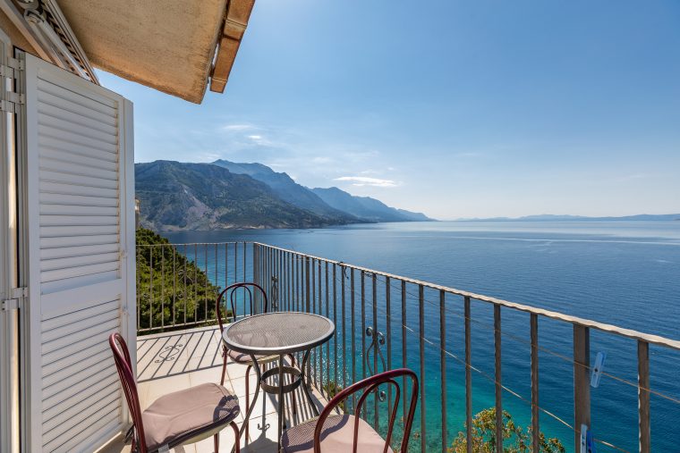 Balkon sa stolom i stolicama te metalnom ogradom, s panoramskim pogledom na plavo more, planinsku obalu i udaljene otoke.