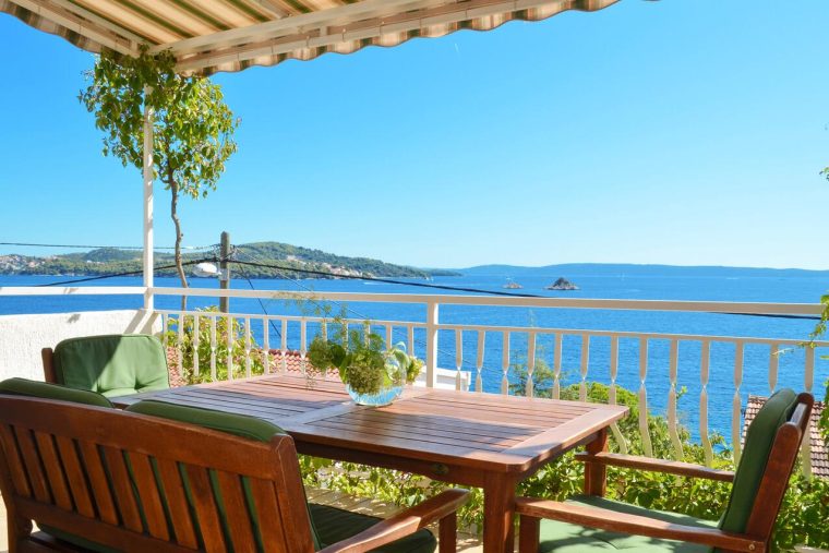 Natkrivena terasa s drvenim stolom i stolicama sa zelenim jastucima, okružena mediteranskim biljem, s panoramskim pogledom na plavo more i obližnje otoke.